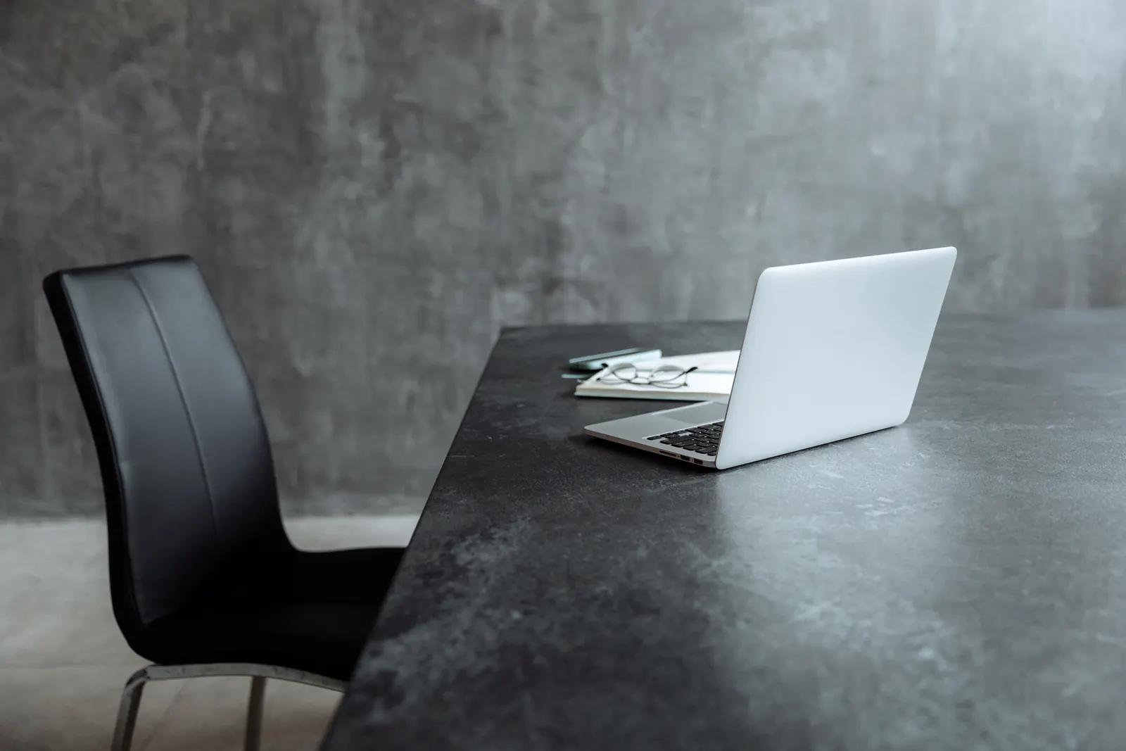 Poste de travail individuel avec ordinateur portable, lunettes et chaise en cuir sombre autour d’une table à la texture minérale.